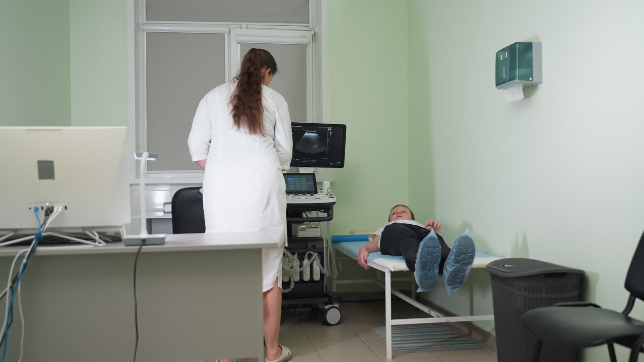 Medical practitioner directs boy to lie on examination bed in lab while drawing chair to ultrasound machine preparing to conduct scan with transducer in clinical setting focused on diagnostics