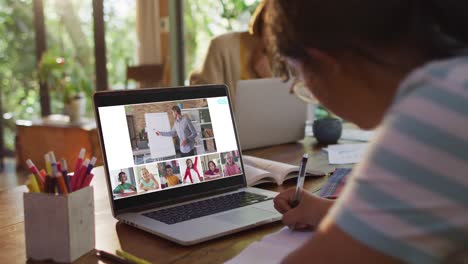 Schoolgirl-using-laptop-for-online-lesson-at-home,-with-diverse-teacher-and-class-on-screen