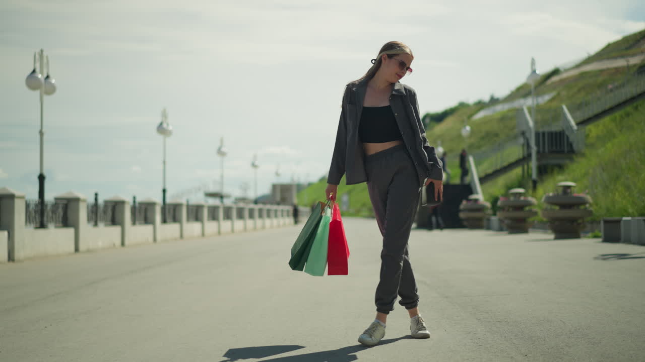 선글라스를 입은 여자가 오른손에 쇼핑백을 들고 행복하게 춤을 추고, 그녀의 그림자는 바닥에 보이고, 한쪽에는 언덕이 있고, 사람들은 배경에 계단 근처에서 고 있습니다.