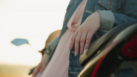 primer plano de la mano de una mujer con uñas pintadas descansando en un asiento de motocicleta, ella lleva una chaqueta de vaqueros y un vestido rosa, con un fondo suavemente borroso