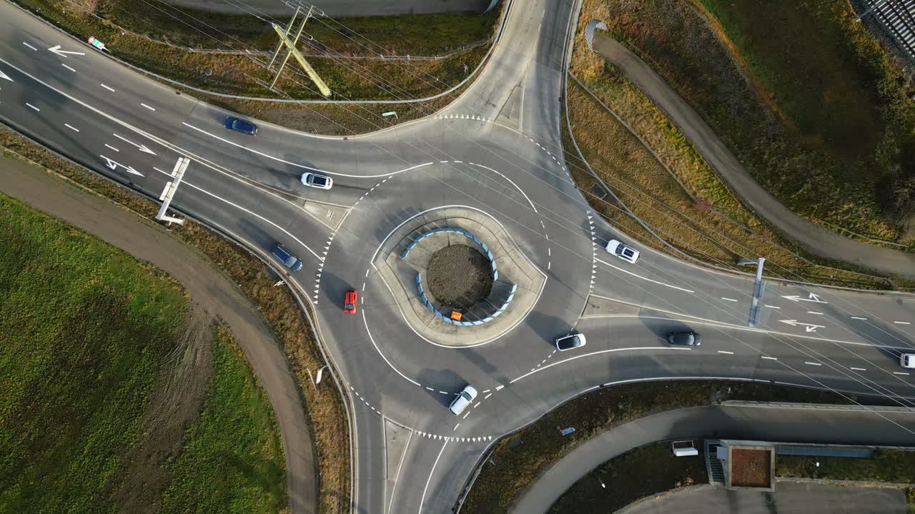 Drone shot looking straight down at a multi-lane roundabout with cars and trucks circulating smoothly, surrounded by rural roads and fields