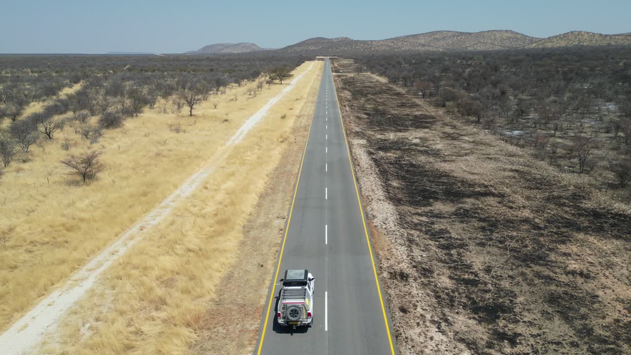 Vista aerea da drone nel deserto della Namibia, con un'auto SUV che percorre una strada stretta, sabbia e alberi verdi ai lati e montagne all'orizzonte in una giornata di sole