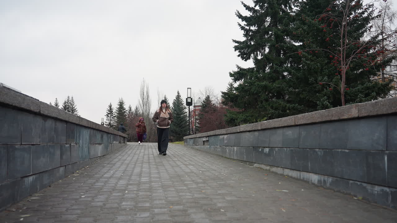 Student wearing black cap brown jacket and trouser walking slowly with hands in pockets on a pathway with pine trees and buildings under overcast sky during cold autumn, another person follows behind