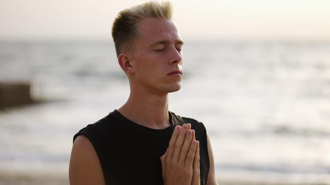 Portrait of a young man meditating against the backdrop of the waves of the sea during dawn. Close-up. Hands near the chest