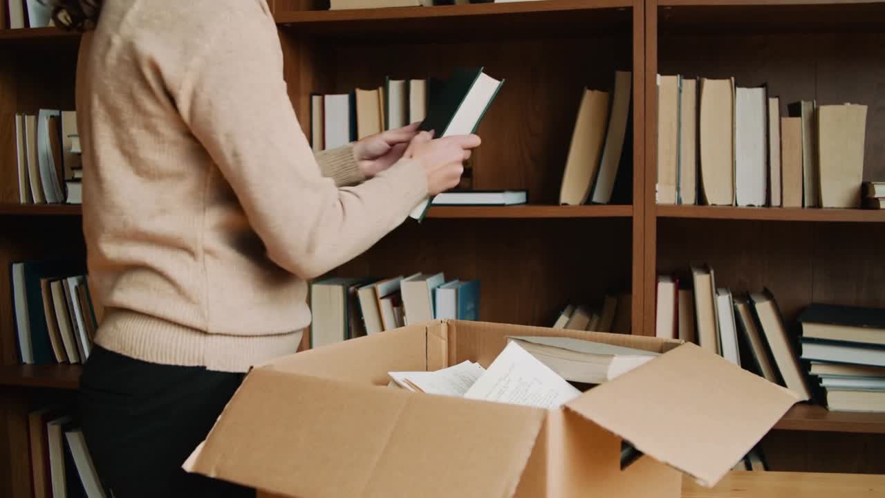 Reaching into box, woman in tan sweater unpacking books in study, shelving green book, copy space