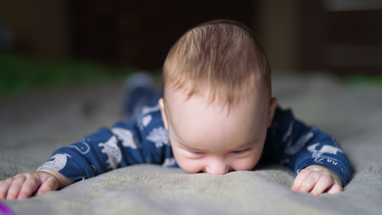 Smiling infant baby boy lying on his belly on bed. Cute boy in blue suit at grey blurred background.