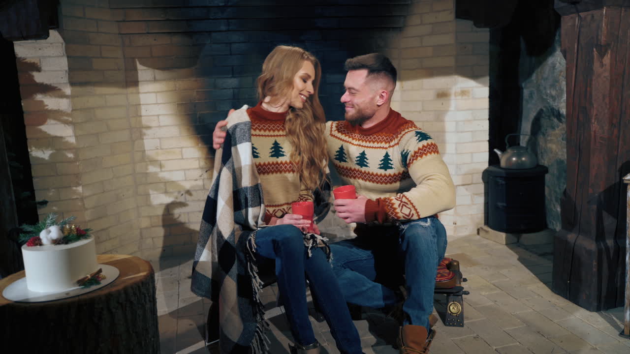 Beautiful couple is sitting near fireplace drinking tea. Man tenderly hugs woman. Intimate atmosphere.