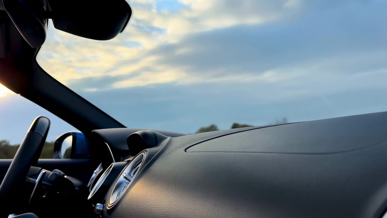 Fixed cabin view shows steering wheel and round air vents beside a broad textured dashboard while changing light and distant trees pass beneath layered blue and golden clouds.
