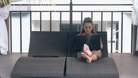 barefoot girl sits with laptop on couch in tropical hotel