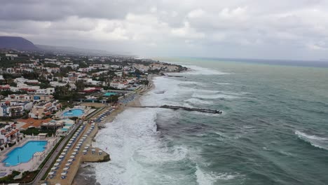 footage of a drone soaring high above the sea, capturing the power of the big waves crashing against the city pier in the background, all set against the stunning backdrop of dark, moody skies.