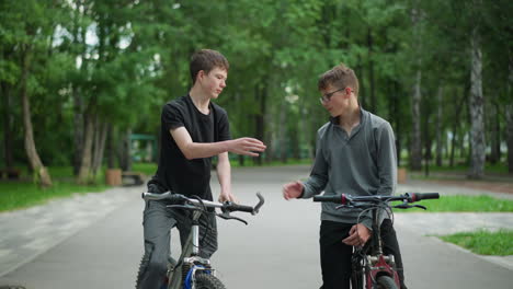 two brothers on bicycles share a friendly moment in a park setting, one boy in a grey top waits while the other rides up to greet him with a knuckle bump, background features lush green trees