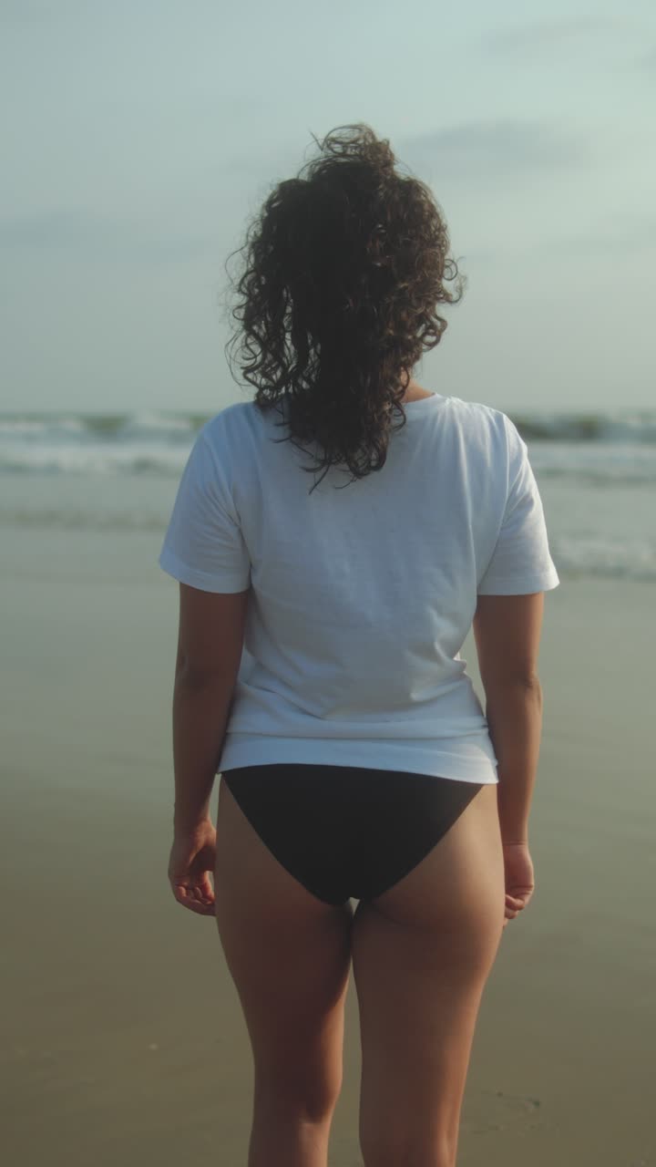 Woman in white t-shirt and black bikini bottoms stands facing ocean waves on sandy beach