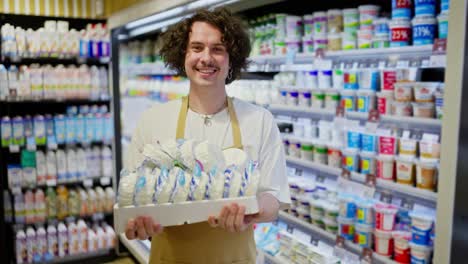 Zoom-Out-portrait-of-a-Happy-guy-with-curly-hair-standing-and-holding-dairy-products-in-his-hands-in-the-department-of-the-supermarket-where-he-works