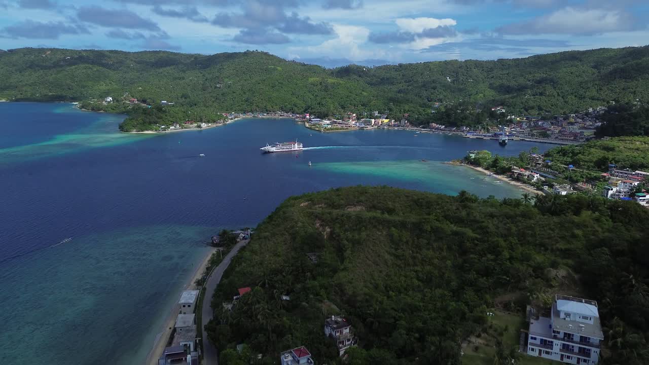 Drone aereo che riprende una grande nave passeggeri in uscita dal porto di Romblon, circondata da acque cristalline color turchese, colline verdi e cielo blu, mostrando un paesaggio costiero e isolano tropicale e suggestivo