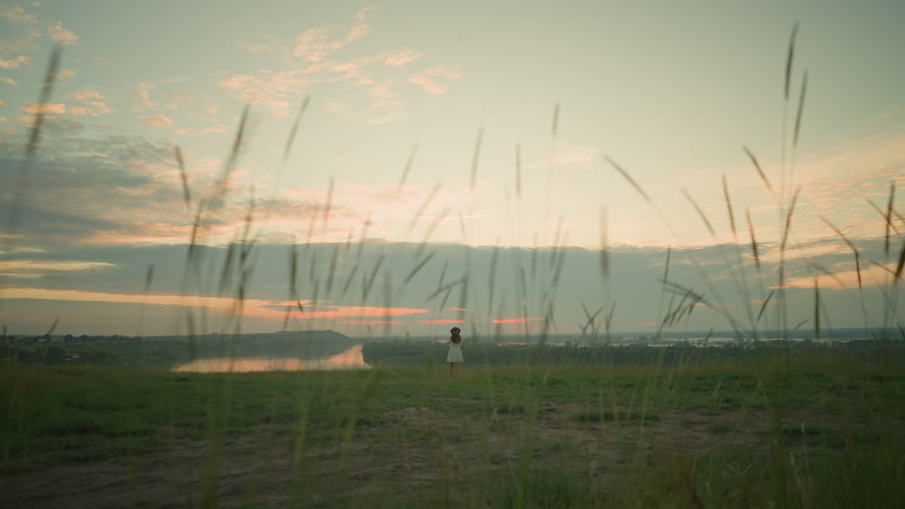 una mujer con un vestido blanco y un sombrero se encuentra sola en un campo de hierba aislado junto a un lago tranquilo al atardecer. ella mira el entorno sereno, abrazando el momento pacífico y reflexivo