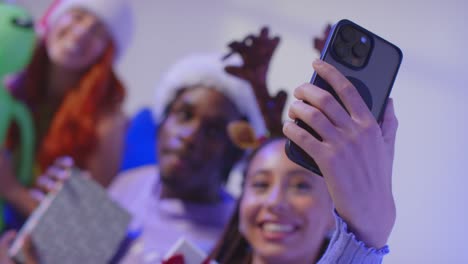 foto in studio di amici della generazione z a natale seduti sul divano con il cappello di babbo natale e le corna di renna che si fanno un selfie con il telefono cellulare 2