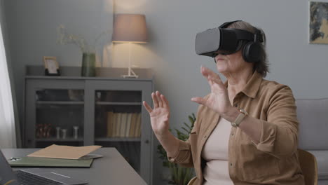 senior woman using virtual reality headset glasses and moving her hands while sitting on chair in a modern living room