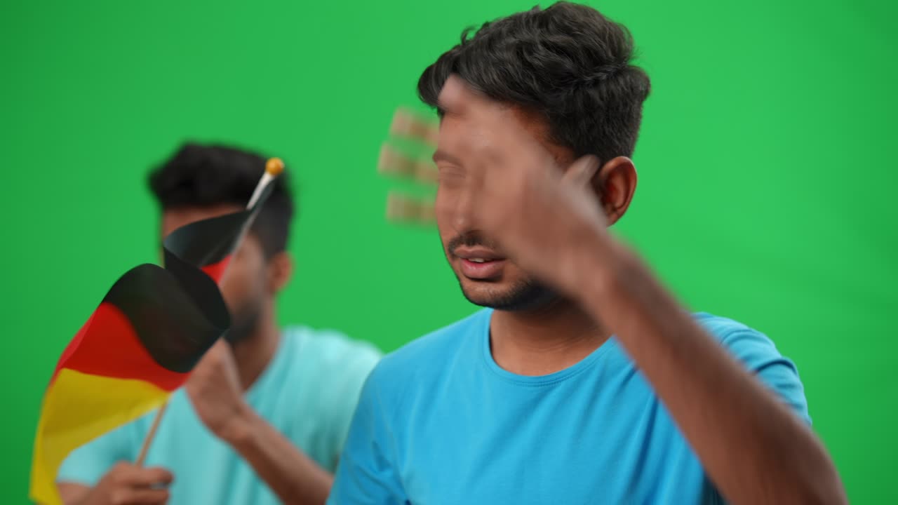 손에 머리를 들고 푸른 스크린에서 독일 발을 들고 흐릿한 친구를 바라보는 스트레스를 받은 축구 팬, 축구 경기를 보는 불만족한 중동 젊은 남자의 초상화.