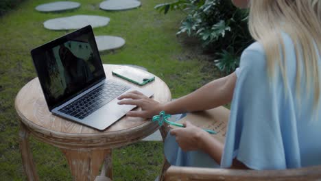 a girl in a blue dress remotely online working behind a laptop and looking into the screen the backyard with green plants