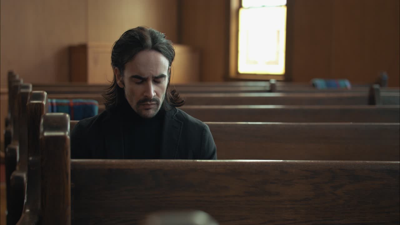 Young man with dark hair, emotional, sombre man in black suit sitting in pew in church in cinematic slow motion
