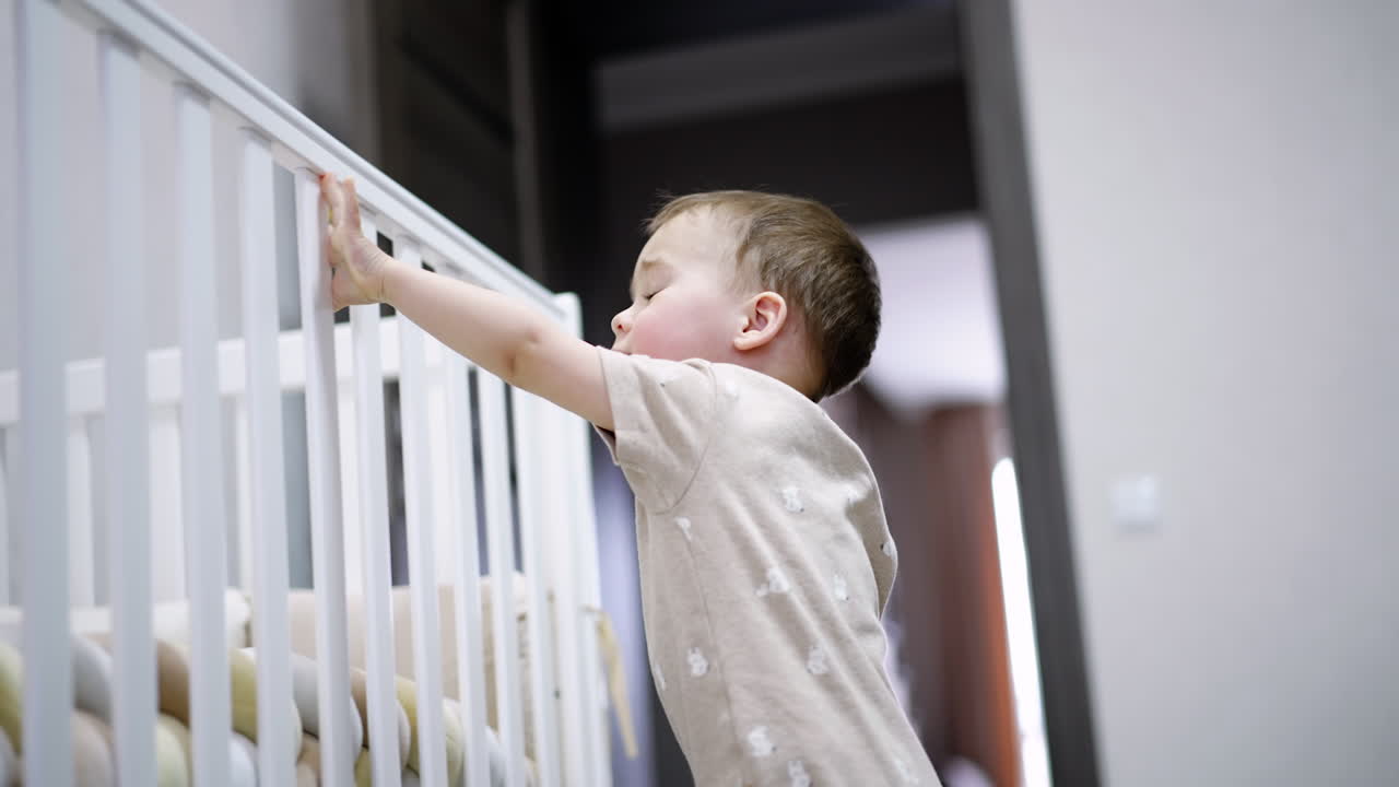 Caucasian boy stands near his white crib. Little child shakes his bed and then watches hoe it moves. Low angle view.