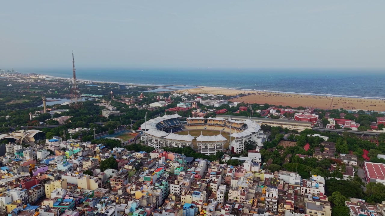 Ripresa aerea con drone che mostra uno stadio di cricket circondato da edifici densi e vegetazione, con Marina Beach e la costa del Golfo del Bengala visibili a Chetpet, Chennai, Tamil Nadu, India.