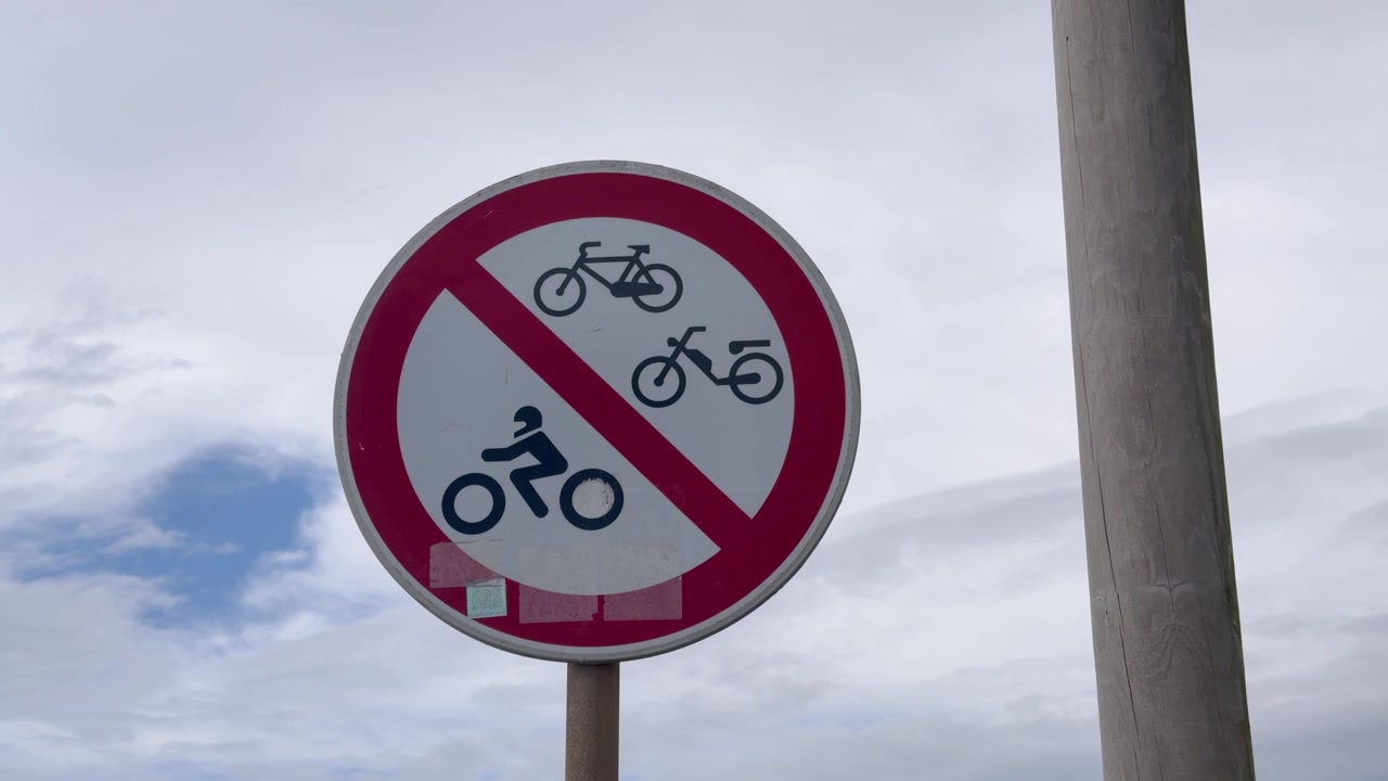 Close up of a red and white sign banning bicycles mopeds and motorcycles under a cloudy sky