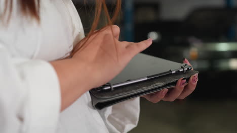 close up of woman in lab coat using tablet in automotive workshop, recording data with well-manicured nails, background blurred, showing industrial setting and advanced technology