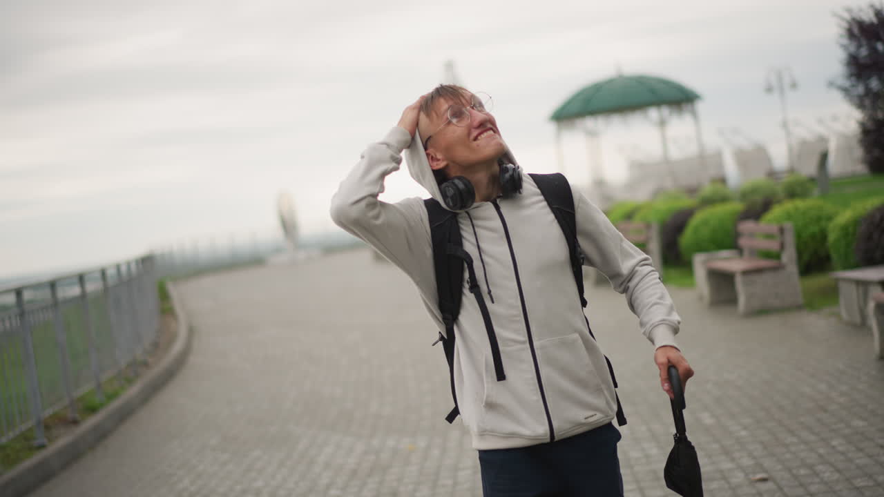 우산을 들고 배낭을 멘 젊은 남자가 비 오는 산책로에서 하늘을 확인하며 물가 난간을 따라 걷고 있어요. 흐린 구름, 젖은 보도, 멀리 보이는 정자, 생각에 잠긴 분위기, 비를 피할 곳을 찾고 있습니다.
