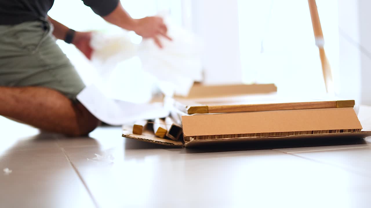 A person unpacks a cardboard box on a sunlit floor, revealing wrapped items. Natural lighting creates a warm atmosphere