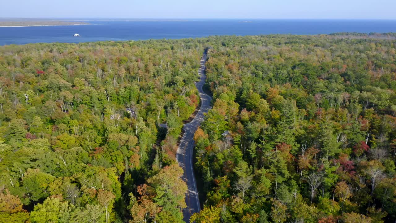 A long winding road threads through a vast forest of early autumn colors, drawing the eye toward a distant blue lake and horizon where land, water, and air meet in calm symmetry