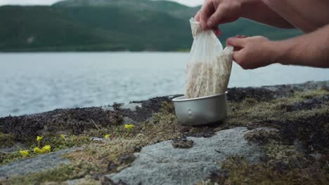 camper pours rice grain into pot for cooking outdoor by the lake shore