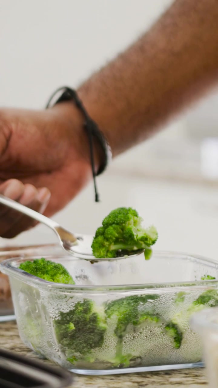 Video verticale: Una mano che prende i broccoli con un cucchiaio e li mette in vassoi neri in cucina per la preparazione.