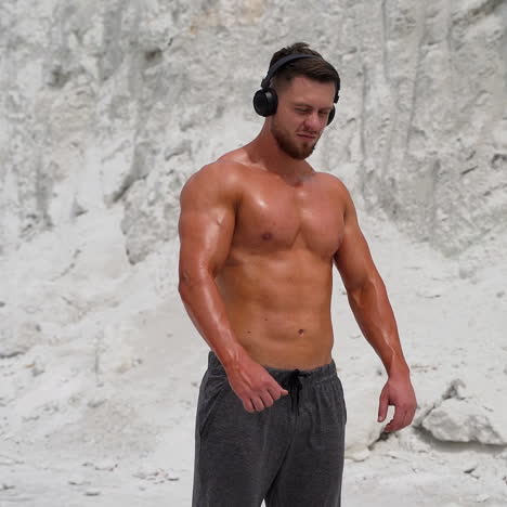 strong man with headphones is doing exercises on the mountain background. young shirtless male with muscular body is training in the open air in white mountains.