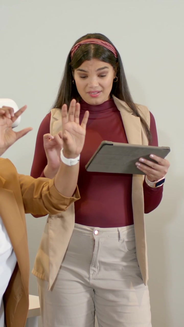 Video verticale: Guardando un tablet, una donna con un gilet che tocca lo schermo e legge in ufficio, mentre un collega conta delle cifre.