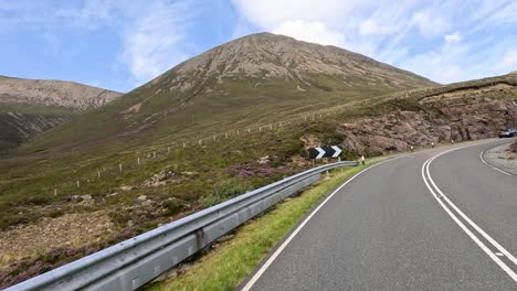 A winding mountain road curves past rocky cliffs and green hills under daylight, captured in a smooth forward-moving perspective with clear, natural lighting