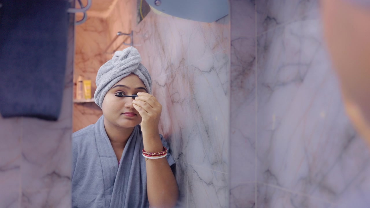 Young lady applying mascara on eyelashes while looking in the mirror, wearing bathrobe in modern bathroom.