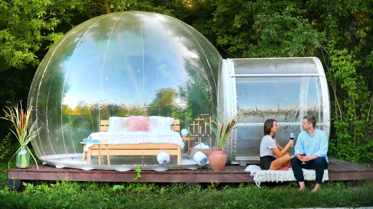 Happy couple drinking wine near transparent bubble tent at glamping. Nature around