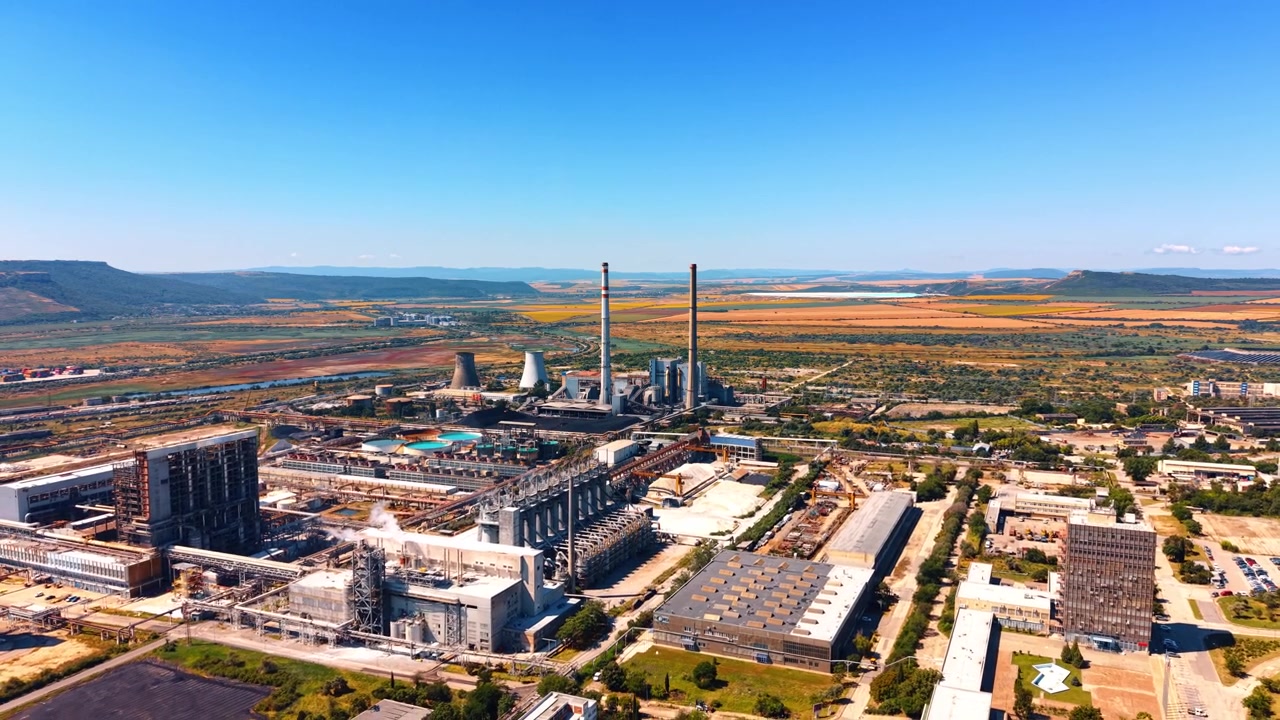 Vista aerea panoramica di un vasto complesso industriale. Edifici industriali, condotte e ciminiere si estendono verso colline lontane sotto un cielo estivo sereno.