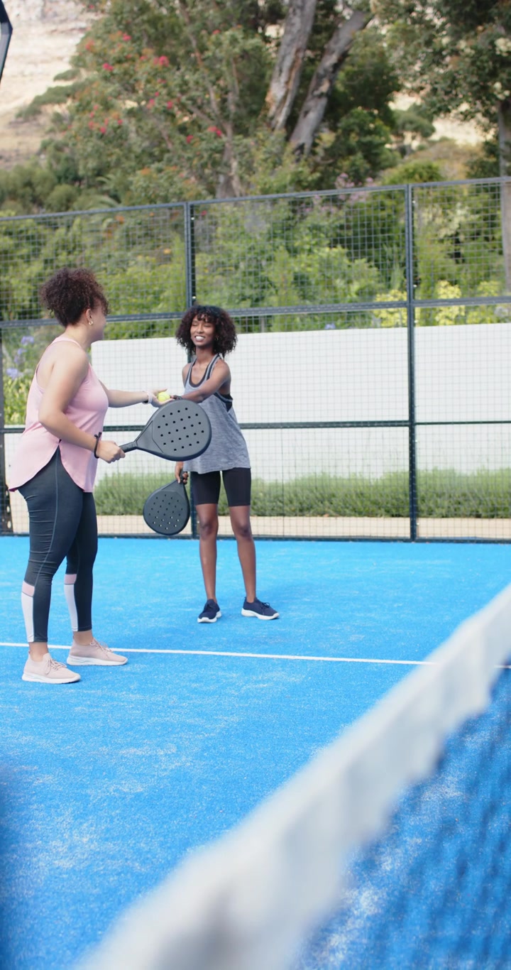 Vertical video: African American women entering blue court, exchanging ball and paddles for warmup
