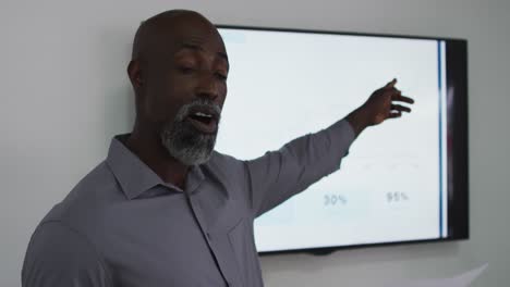 African-american-businessman-talking-and-pointing-at-screen,-giving-presentation-in-office