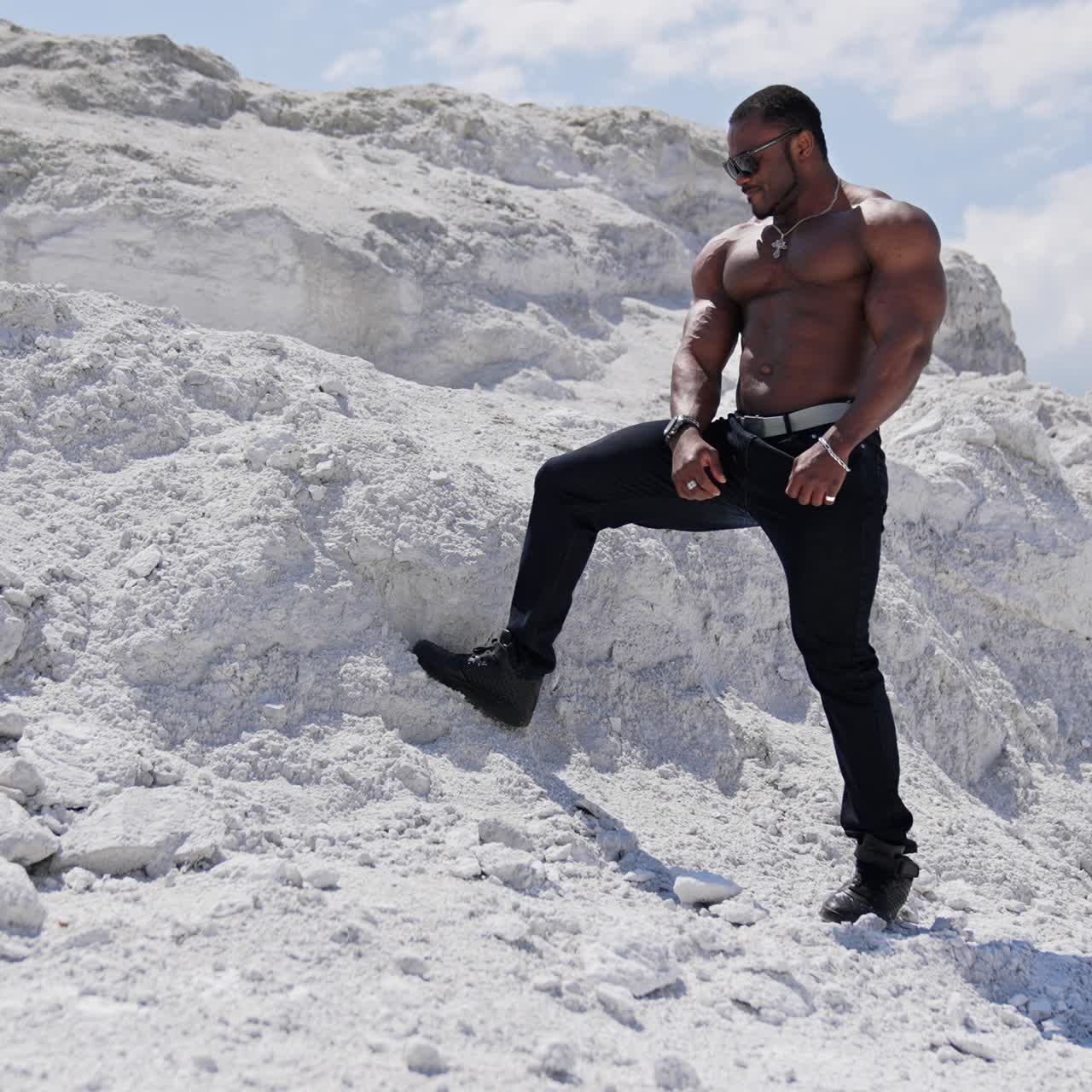 Handsome bodybuilder posing on camera. Sportive african american man in black glasses without shirt showing his strong muscles against white hill