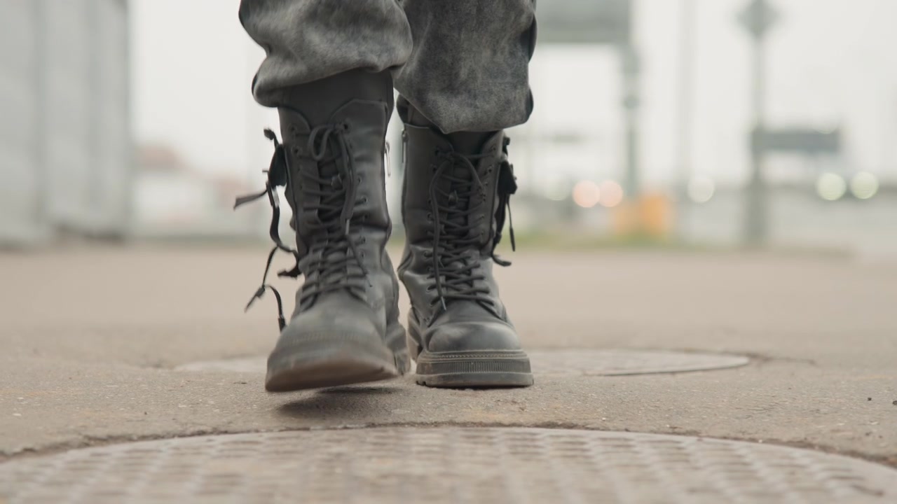Boots stepping on manhole cover in street, lowangle closeup showing scuffed leather, laces and purposeful pace, muted urban background, gritty determined motion ideal for journey or military themes