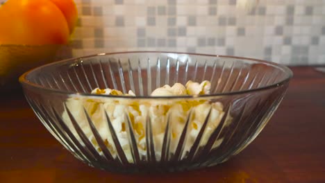 Close-up-slow-motion-footage-of-popcorn-being-poured-into-a-glass-bowl-on-a-kitchen-table-with-oranges-and-lemons-in-a-bowl-in-the-background