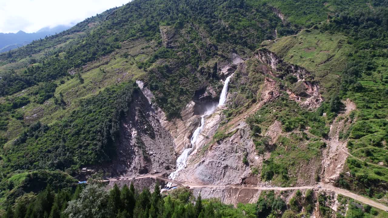 drone volando más allá del pico de la montaña y revela rupees cae cascada y coches conduciendo en la carretera de tierra de la montaña verde en nepal