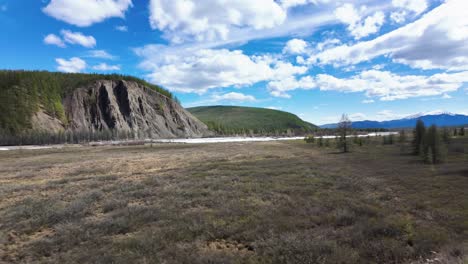 A-vast,-open-field-with-a-river-running-through-it,-surrounded-by-mountains-and-trees-under-a-bright-blue-sky