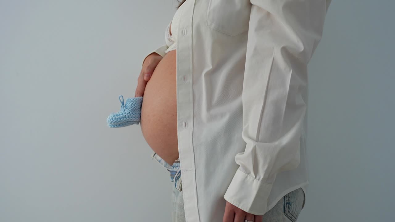 Pregnant woman holding blue baby booties in front of her belly, zoom out shot