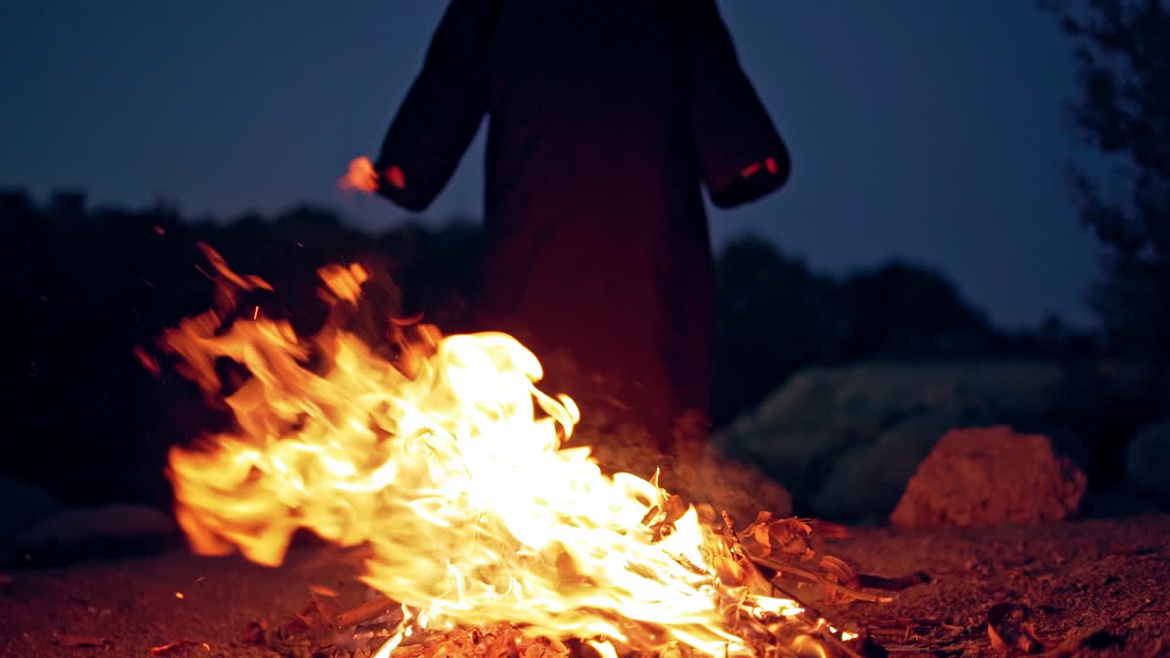 Fire explodes on the ground before silhouette of death. Black robe waving arms near the bright flame in the evening. Horror ghost at night.