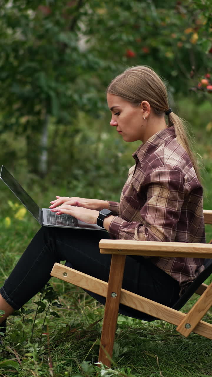 Blonde young lady typing on her laptop sitting in the apple garden. Woman working freelance in the nature surrounding. Vertical video