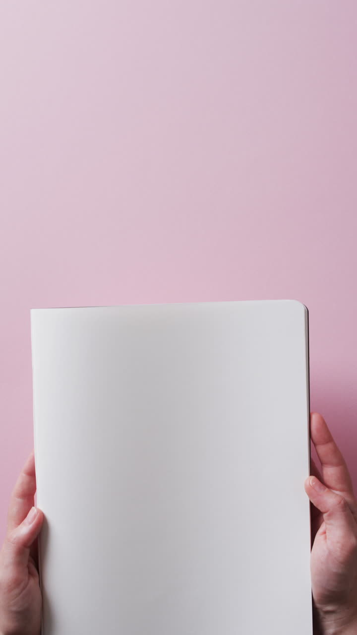 Vertical video of hand holding book with copy space on pink background in slow motion
